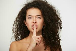 Emotional young girl with curly hair, calls for silence, do not tell anyone,keep quiet, do not make noise, keeps fore finger on lips,demonstrates silence gesture,head and shoulders,on white background photo