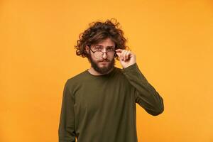 Tthe young professor lowers his glasses with interest and examines something new. Bearded curly-headed man looked something curious and takes off his glasses photo