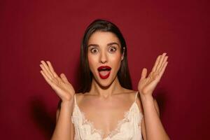 Astonished young pretty brunette lady with evening makeup rounding her brown eyes while looking surprisedly at camera with raised hands, standing over burgundy background photo