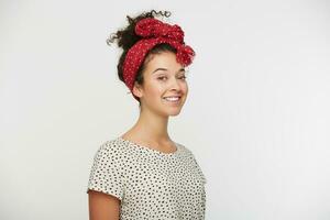 Headshot of tender curly hair woman with beautiful eyes, standing in a half-turn charmingly smiling, feels happiness, shows tenderness, wears white shirt with black polka dots. Over white background photo