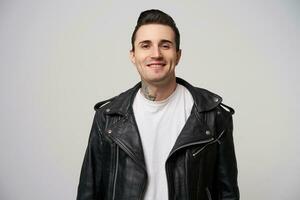 Charmingly, enticing, boldly smiling young rocker, rebel, motto racer with a stylish hairstyle in briolin, tattoo on the neck, dressed in the style of 50s-60s in black leather jacket, white background photo