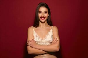 Charming cheerful young long haired brunette woman with festive makeup crossing her arms on chest and looking happily at camera with wide smile, isolated over burgundy background photo