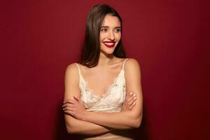 Studio shot of lovely young dark haired female with evening hairdo and red lips folding her hands on chest and looking cheerfully aside with charming smile, posing over burgundy background photo