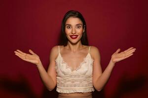 Studio photo of young brown-eyed brunette lady with festive hairstyle shrugging with raised palms and looking suprirsedly at camera, isolated over burgundy background