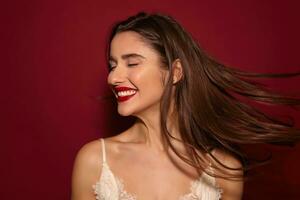 Pleasant looking young attractive happy brunette lady with red lips turning her head and laughing cheerfully with closed eyes, isolated over burgundy background photo