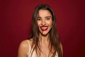 Indoor photo of young attractive dark haired female with evening makeup and wild hairstyle looking cheerfully at camera with broad smile, posing over burgundy background