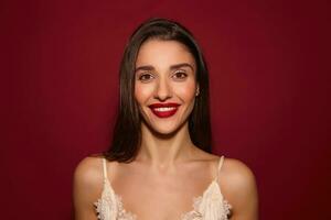 Portrait of young cheerful beautiful dark haired female with festive mua looking positively at camera with wide charming smile, standing over burgundy background photo