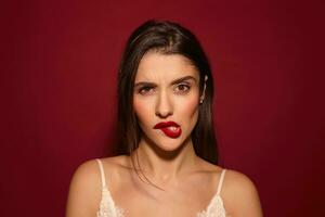 Portrait of young brown-eyed pretty dark haired lady with festive makeup looking seriously at camera and biting underlip, posing over burgundy background photo