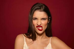 Displeased young attractive brunette female with casual hairstyle frowning her face and biting lip while looking at camera, standing over burgundy background photo