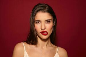 Indoor photo of serious young brunette female with festive hairstyle twisting her mouth while looking at camera, wearing white strap top while posing over burgundy background