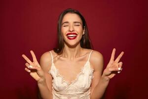 Good looking joyful young beautiful dark haired lady with evening makeup raising piece gesture with both hands and smiling broadly with closed eyes, isolated over burgundy background photo