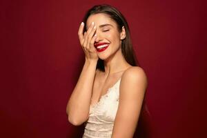 Positive young charming long haired brunette woman keeping her eyes closed while smiling cheerfully and holding raised palm on her face, isolated over burgundy background photo