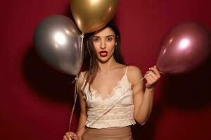 Studio shot of young brown-eyed long haired brunette woman with evening makeup holding air balloons and looking seriously at camera, isolated over burgundy background photo