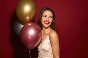 Indoor shot of young attractive dark haired female with evening hairstyle standing over burgundy background with multi-colored helium balloons and smiling widely photo