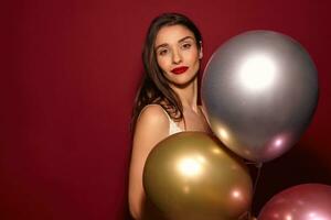 Charming young brown-eyed dark haired female with festive makeup looking calmly at camera and smiling slightly, standing over burgundy background with multi-colored helium balloons photo