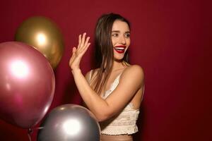 Cheerful young lovely brunette woman dressed in white strap posing with air balloons over burgundy background, being in high spirit while celebrating something photo