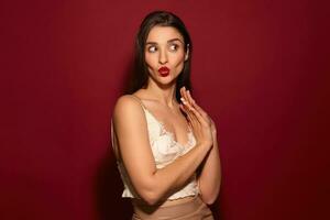 Studio shot of young lovely dark haired female with festive makeup pursing her lips and looking excitedly aside, keeping raised hands folded while standing over burgundy background photo