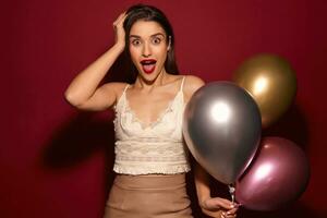 Beautiful young dark hairedb elegant lady being surprised by friends on her birthday, looking amazedly at camera with wide eyes and mouth opened, isolated over burgundy background photo