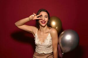 Glad young attractive long haired brunette female with festive makeup keeping victory gesture near her face and looking cheerfully at camera, isolated over burgundy background photo