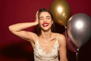 sorprendido joven atractivo oscuro peludo hembra con noche maquillaje sonriente extensamente con cerrado ojos y participación elevado mano en su cabeza, alegría mientras celebrando alguna cosa terminado borgoña antecedentes foto