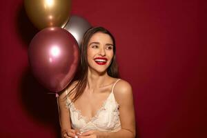 Happy young elegant long haired brunette pretty woman looking cheerfully aside and smiling sincerely, rejoicing about nice party with her friends while posing over burgundy background photo