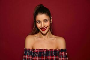 Indoor shot of young beautiful brown-eyed cheerful brunette lady looking at camera with charming smile while posing over burgundy background with naked shoulders photo