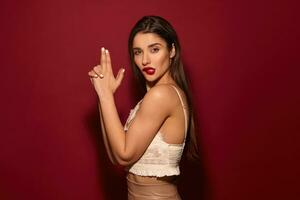 Charming young brown-eyed long haired brunette woman with evening makeup folding gun with raised hands and biting underlip while looking at camera, posing over burgundy background photo