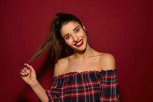 Happy young beautiful long haired brunette woman with ponytail hairstyle looking gladly at camera and smiling pleasantly, dressed in checkered top while posing over burgundy background photo