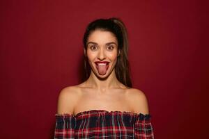 Funny young attractive brown-eyed brunette female with casual hairstyle fooling and sticking cheerfully out her tongue while looking at camera with excited face, isolated over burgundy background photo