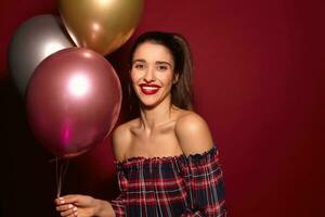 Joyful young beautiful dark haired female with casual hairstyle holding bunch of multi-colored helium balloons and smiling happily at camera, isolated over burgundy background photo