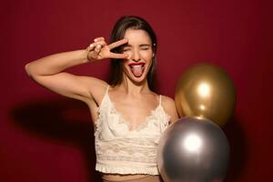 Portrait of young cheerful brunette female with evening makeup fooling while posing over burgundy background with air baloons, raising victory sign to her face and showing tongue photo