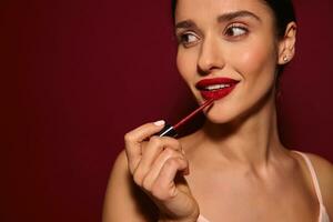 Close-up of attractive young brunette female with ponytail hairstyle applying lip gloss on her lips and looking aside with charming smile while standing over burgundy background photo