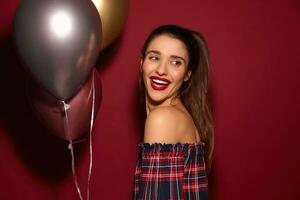 Indoor photo of cheerful young lovely long haired brunette lady with ponytail hairstyle having fun on party and laughing happily while looking aside, isolated over burgundy background