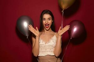 Astonished young beautiful brunette female with evening makeup raising emotionally her palms and looking at camera with wide eyes and mouth opened, isolated over burgundy background photo