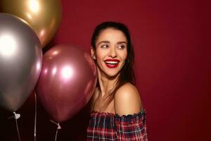 Happy young pretty dark haired woman with festive makeup looking cheerfully aside with charming smile while posing over burgundy background with air balloons photo