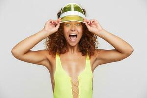 Bemused young pretty red haired woman with casual hairstyle raising emotionally hands to her cap while looking at camera with wide eyes and mouth opened, posing over white background photo