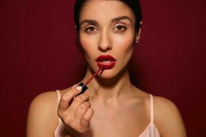 Studio photo of young pretty brown-eyed dark haired female looking seriously at camera while applying lip gloss on her lips, isolated over burgundy background