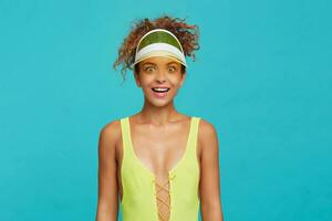 Agitated young pretty red haired curly female rounding surprisedly her green eyes while looking cheerfully at camera, standing over blue background with hands down photo