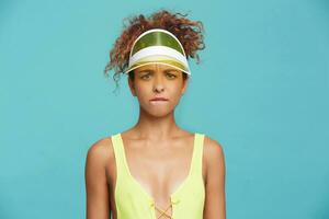 Puzzled young green-eyed redhead curly lady frowning her eyebrows and biting wrringly lips while looking at camera, standing over blue background with hands down photo