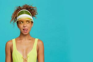 Studio photo of young concerned red haired woman showing her tongue while looking wonderingly upwards, dressed in yellow swimming wear while posing over blue background