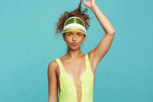 Portrait of young puzzled redhead curly lady with casual hairstyle pulling her hair with raised hand and grimacing confusedly face while posing over blue background photo