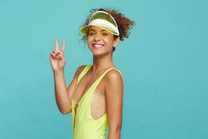 Joyful young attractive red haired curly woman with natural makeup raising hand with victory gesture and smiling widely at camera while posing over blue background photo
