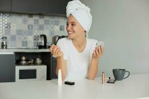 Portrait of a beautiful girl making make-up in the morning, drinking tea, looking and smiling to someone a side, dressed casually and head wrapped in a towel, on a kitchen background photo