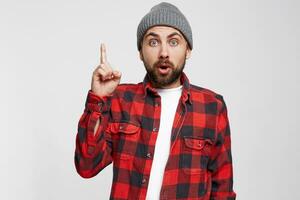 There is an idea, a solution is found, a good thought has come. Eyebrows raised, eyes rounded, shows a finger up. A young attractive bearded guy is wearing a plaid shirt and gray hat,white background photo