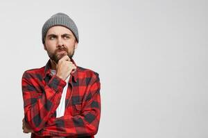 Young european man thinks, raised hand to face, touches his beard, looks dissatisfied, puzzled, in thought, one corner of the lips pursed, white background, blank copy space for text or advertisement photo