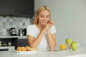 Young blonde girl sits at the kitchen table in the morning, smiling, looks at tablet black screen, on the table -a glass of juice, apples, buns, dressed in a domestic white T-shirt, kitchen background photo