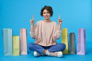 Displeased young short haired brunette lady dressed in pink sweatshirt, jeans and sneakers while posing over blue background, pointing upwards and frowning her face while looking confusedly at camera photo