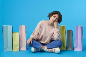 Melancholy young lovely curly brunette female dressed in pink sweatshirt and jeans frowning eyebrows and folding sadly lips while looking upwards, isolated over blue background photo
