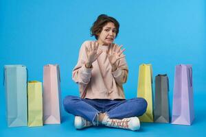 Scared young pretty dark haired curly female raising palms in protective gesture and looking at camera with afraid face, posing over blue background with purchases photo