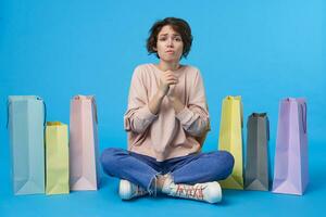 Indoor photo of young dark haired curly lady looking worringly at camera and folding raised hands in praying gesture, posing over blue background with many paper bags